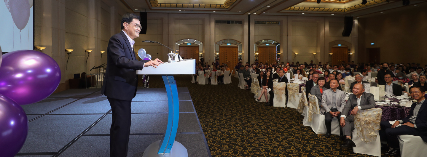Man in suit at podium addresses seated crowd in ballroom, with purple balloons visible.